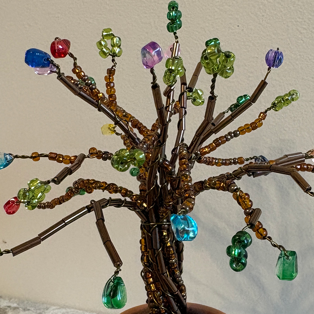 A handmade beaded tree of life wall hanging with brown wire branches adorned with colorful faceted beads in green, blue, red, purple, and clear, shown against a plain beige wall.