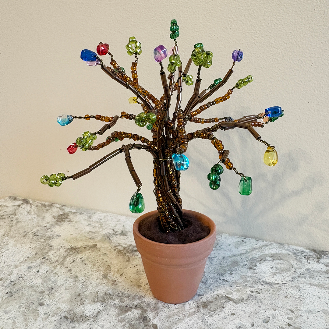 A decorative wire tree of life craft with colorful gem-like beads hanging from brown branches, displayed in a terracotta pot on a stone surface against a plain wall.
