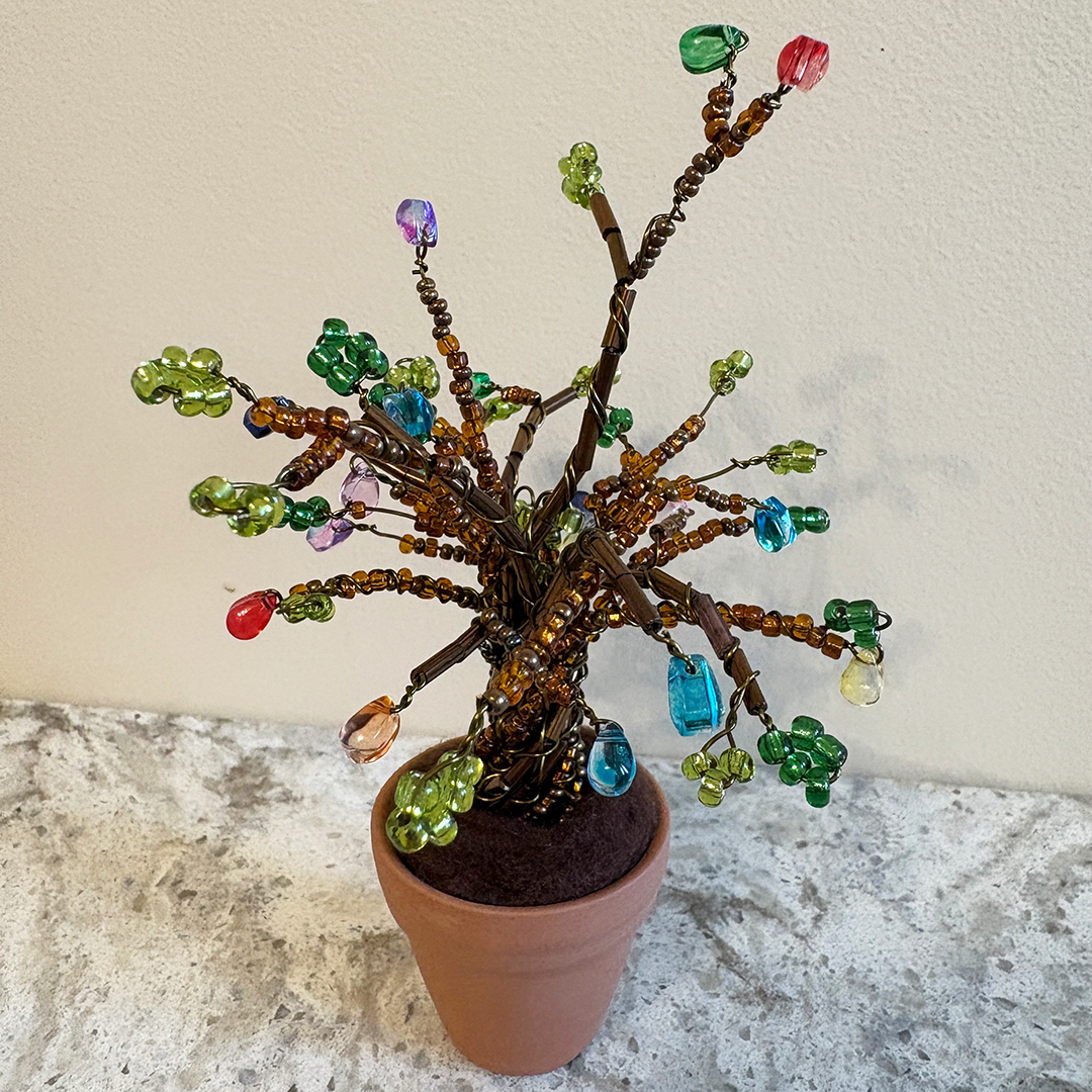 A decorative wire tree of life sculpture with colorful glass beads and charms, displayed in a terracotta pot on a stone surface against a textured beige wall.