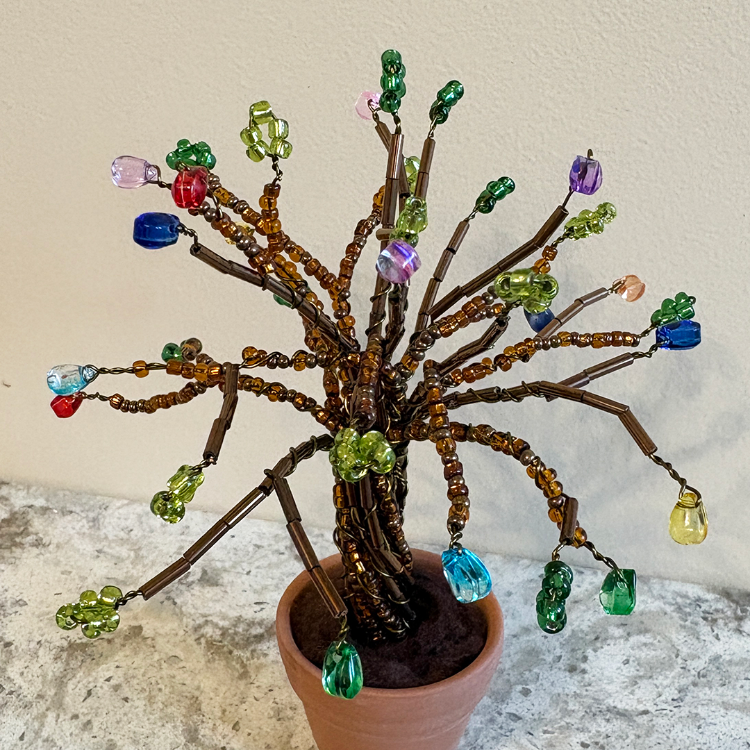 A decorative beaded tree of life figurine with brown twig branches, amber beads, and colorful faceted glass beads, displayed in a terracotta pot against a light beige wall.